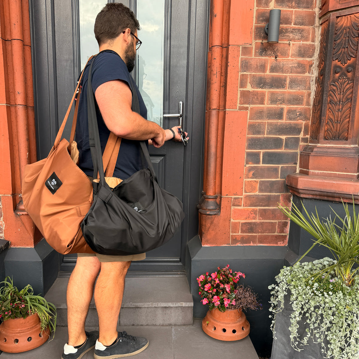 Man entering a house carrying black and cinnamon brown OYSTER lay-flat tote bags filled with shopping.