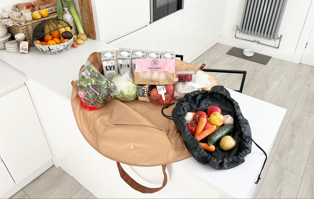 OYSTER lay-flat tote packed with groceries beside an URCHIN drawstring pouch holding fresh vegetables on a kitchen table.