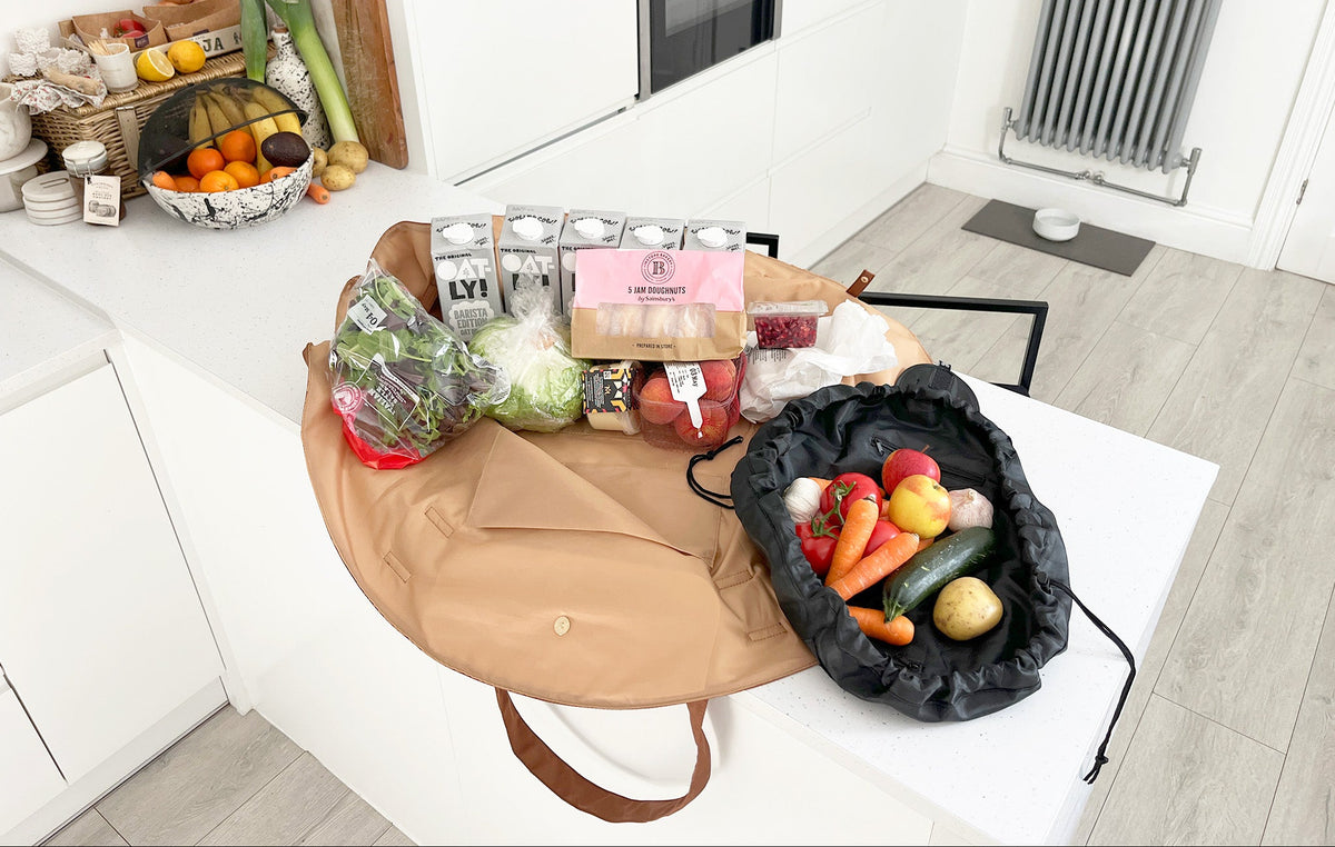 OYSTER lay-flat tote packed with groceries beside an URCHIN drawstring pouch holding fresh vegetables on a kitchen table.