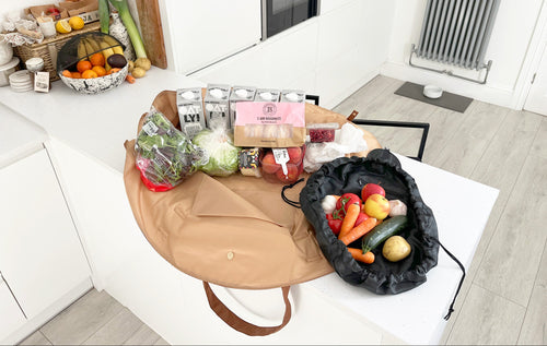 OYSTER lay-flat tote packed with groceries beside an URCHIN drawstring pouch holding fresh vegetables on a kitchen table.