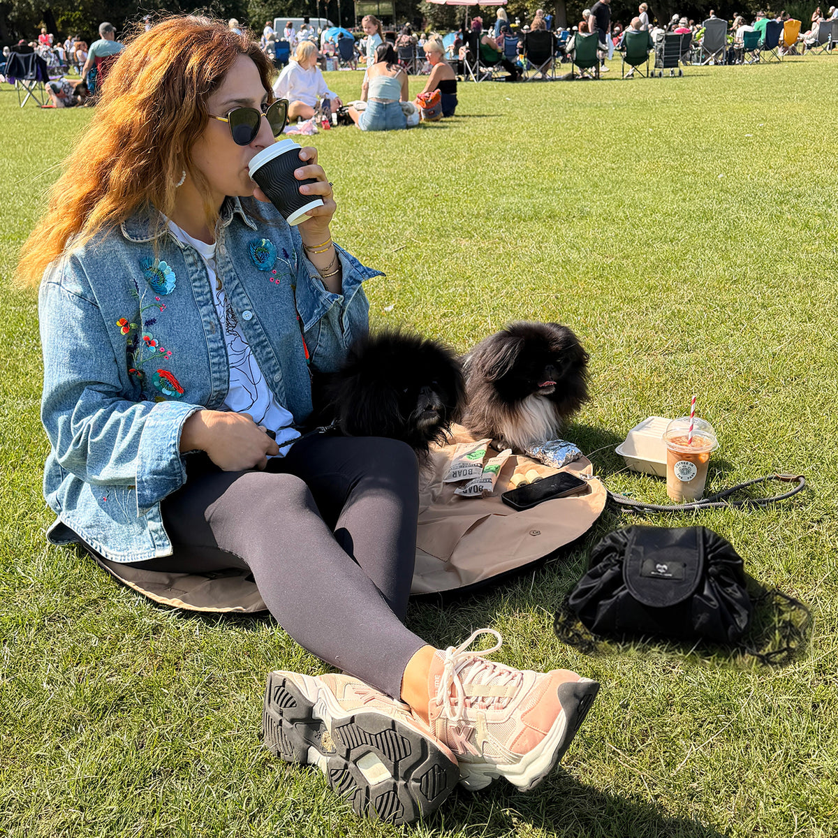 Woman sitting on a cinnamon OYSTER lay-flat tote beside a black URCHIN drawstring pouch at an outdoor park event.