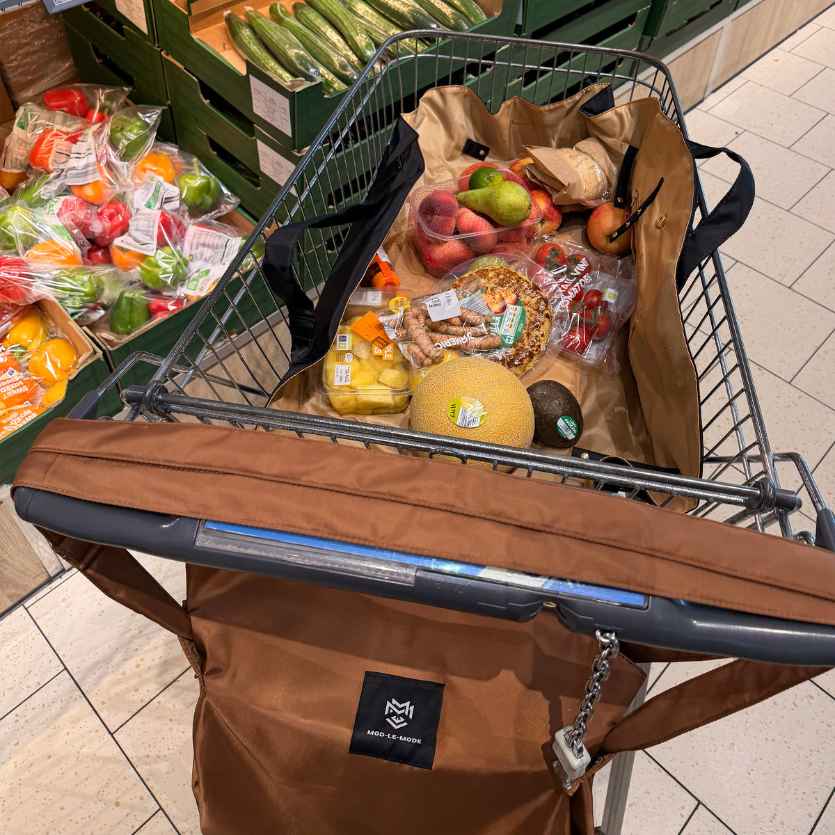 A brown reusable shopping tote bag and a lay-flat tote lines the metal grocery cart in a supermarket. The tote bag has a black logo patch reading 'MOD-LE-MODE' 
